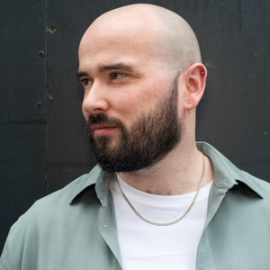 A bald man with a full dark beard and light complexion looks to his left. He wears a light green collared shirt over a white t-shirt, a silver chain, and ear piercings.
