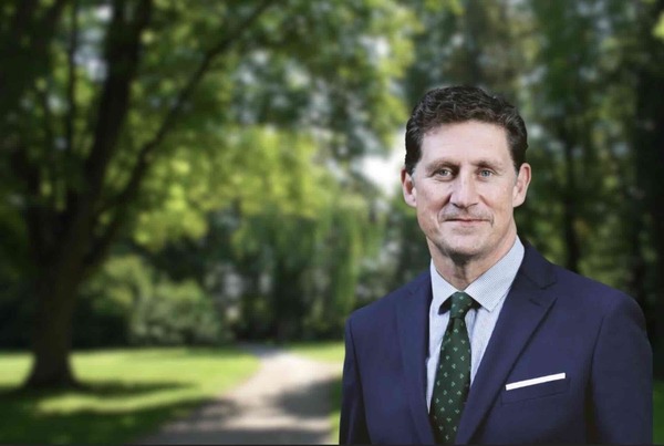 A man with dark hair, blue eyes, and a smile wears a navy suit, light blue patterned shirt, and a dark green patterned tie. He stands outdoors with a blurred green, tree-lined path behind him.