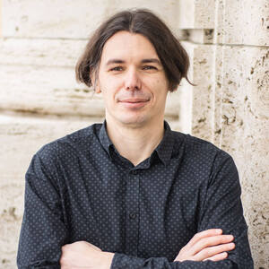 A man with medium dark hair and a subtle smile wears a dark grey shirt with a small geometric pattern, standing with arms crossed against a textured light stone wall.