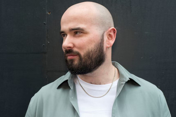 A bald man with a dark beard and two left ear piercings smiles slightly, looking right. He wears a light sage green shirt over a white t-shirt and a gold chain.