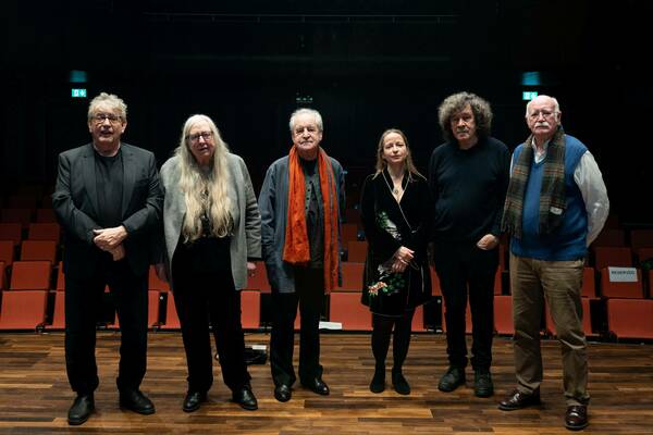 Four men and two women stand on a wooden stage before rows of red theater seats. They include a man in a black suit, a woman with long gray hair, a man with an orange scarf, a woman in an embroidered black robe, a man with curly hair, and a man in a blue vest with a plaid scarf.