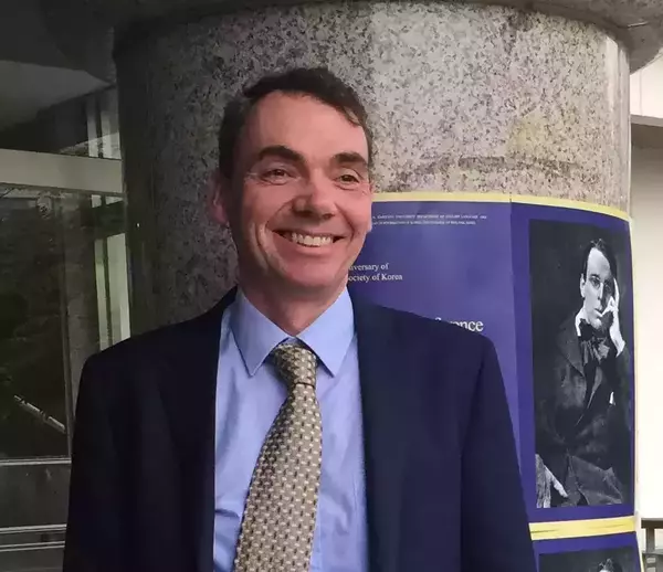 P.J. Mathews in a blue suit and patterned tie stands against a purple banner celebrating an anniversary, featuring an old portrait.