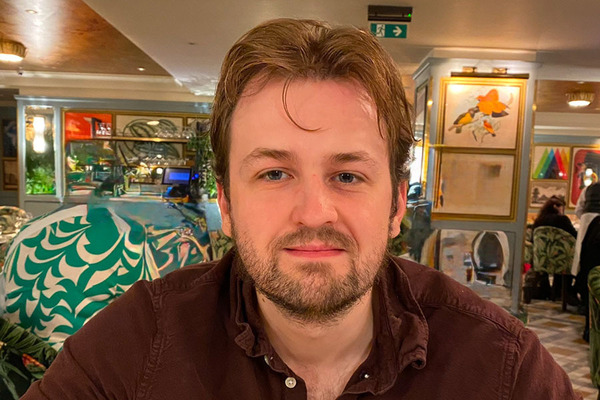 A man with light brown hair, blue eyes, and a beard, wearing a maroon collared shirt, smiles slightly in a restaurant.