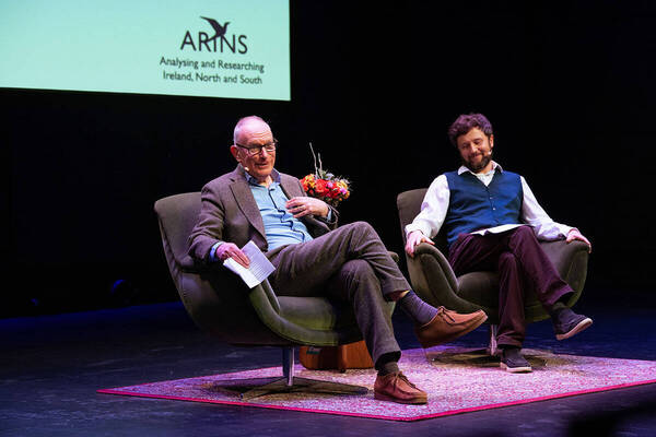 Two smiling men on a stage with an "ARINS" screen. An older man in a brown suit holds papers. A bearded man wears a blue vest.