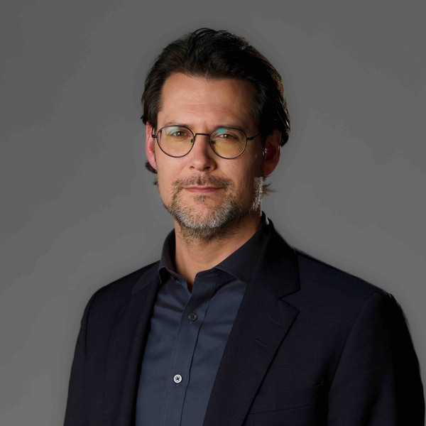 Matt Gertken with dark hair, goatee, and round glasses. He wears a dark blue suit jacket over a dark blue shirt.