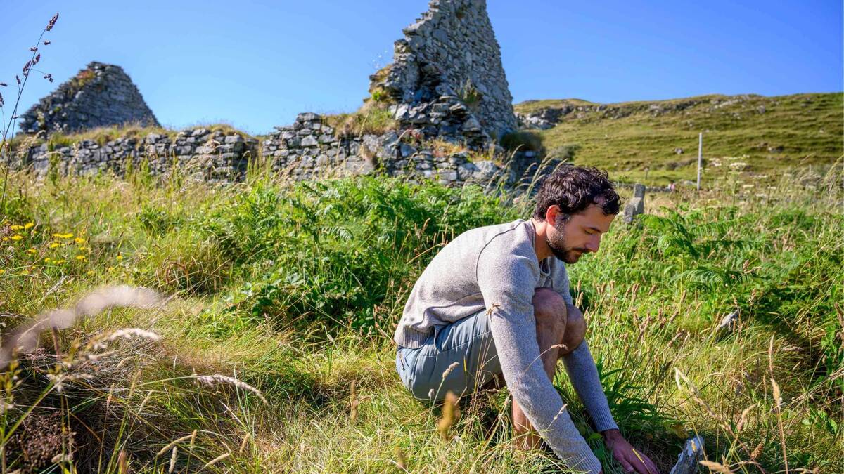 A man in a grey sweater and blue shorts crouches in tall green grass, looking down, with ancient stone ruins behind him.
