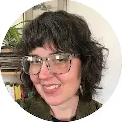 A smiling woman with dark curly hair and bangs wears patterned glasses and an olive corduroy jacket.