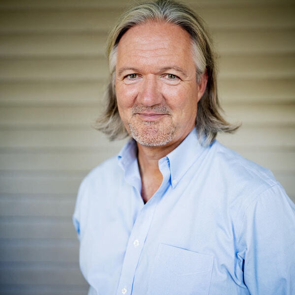 A man with long gray hair and a goatee smiles, wearing a light blue button-up shirt.
