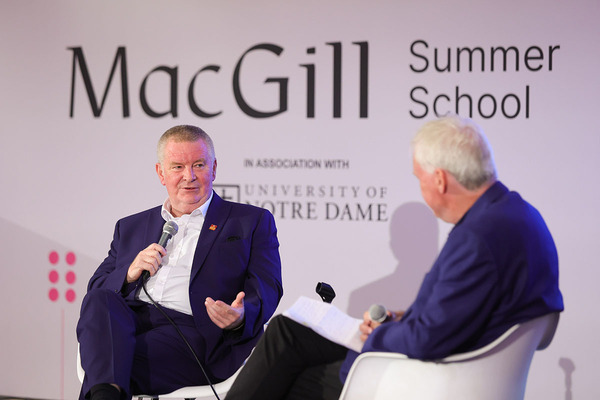 Man in a purple suit speaks into microphone, gesturing, at MacGill Summer School, in association with University of Notre Dame.