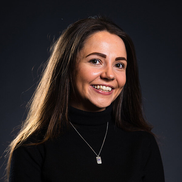 Woman in black turtleneck smiles, wearing a silver necklace with a rectangular pendant.